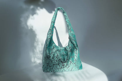 Große Crossbody Tasche in hellblauem Muster und silbernen Reißverschluss steht vor weißem Hintergrund. 