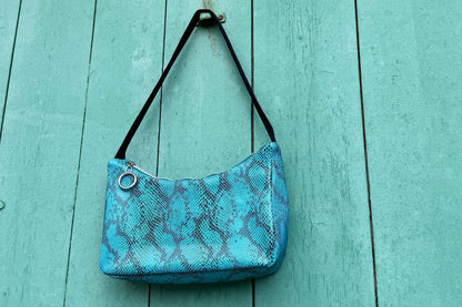 Gesamtansicht blaue Schultertasche in Schlangeleder Optik vor blauem Hintergrund mit schwarzem Gurtband. Hergestellt aus upcycled Materialien von Jeff Every Piece A Friend.