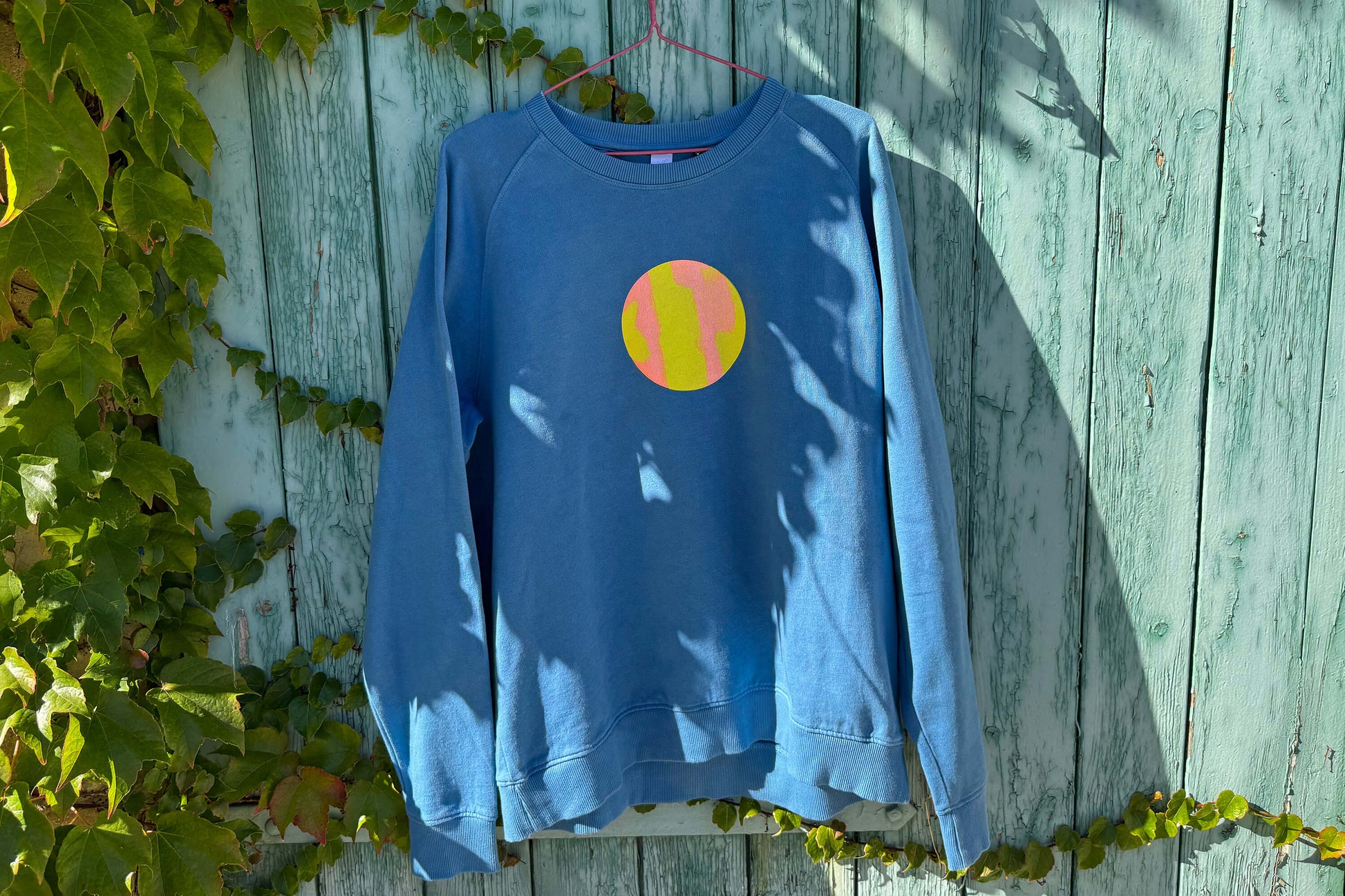 Frontansicht von blauem Sweatshirt vor blauem Hintergrund. Der Pulli hat einen aufgedruckten Kreis in gelb-orange.