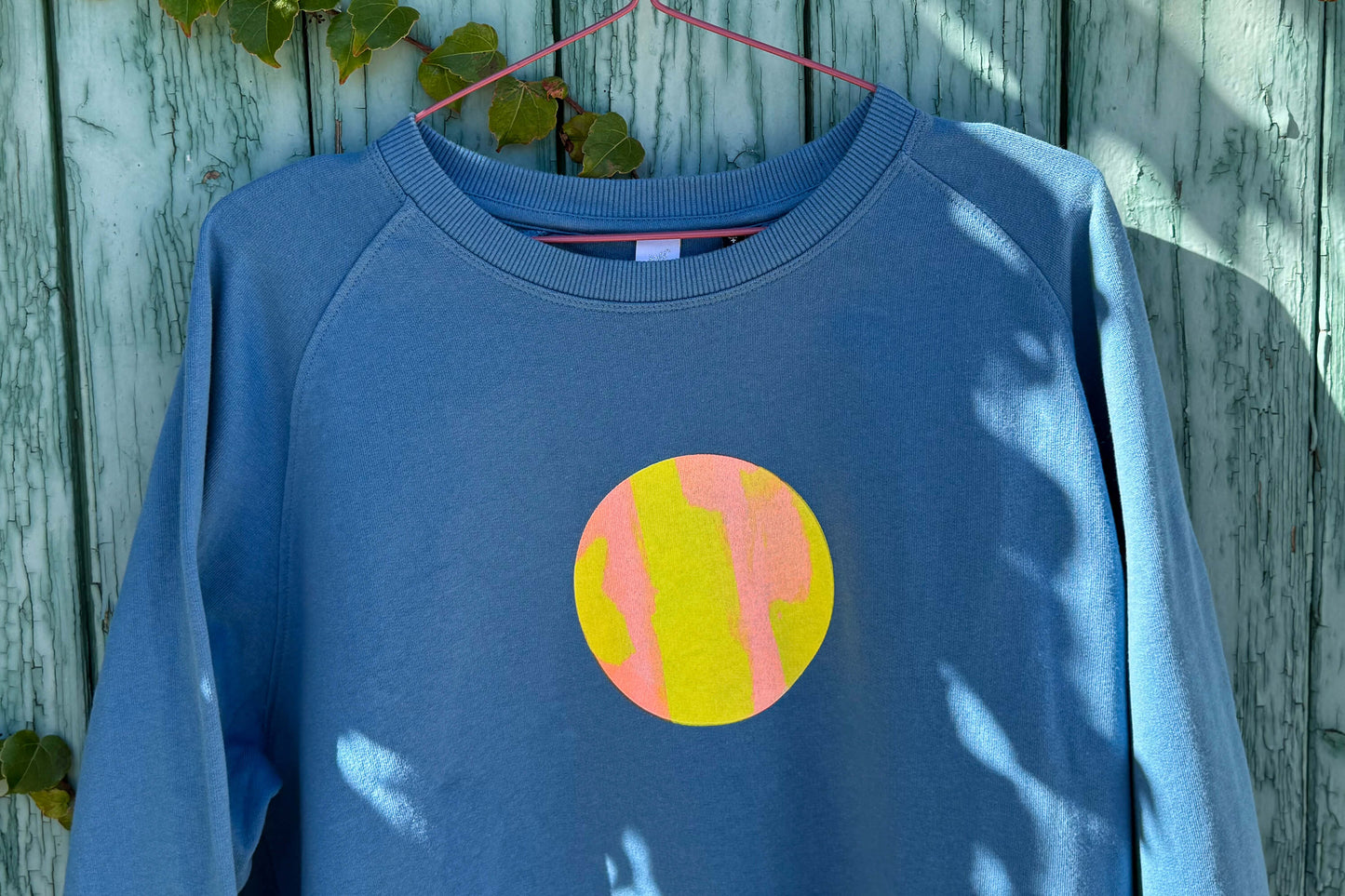 Detailansicht von blauem Sweatshirt vor blauem Hintergrund. Der Pulli hat einen aufgedruckten Kreis in gelb-orange.