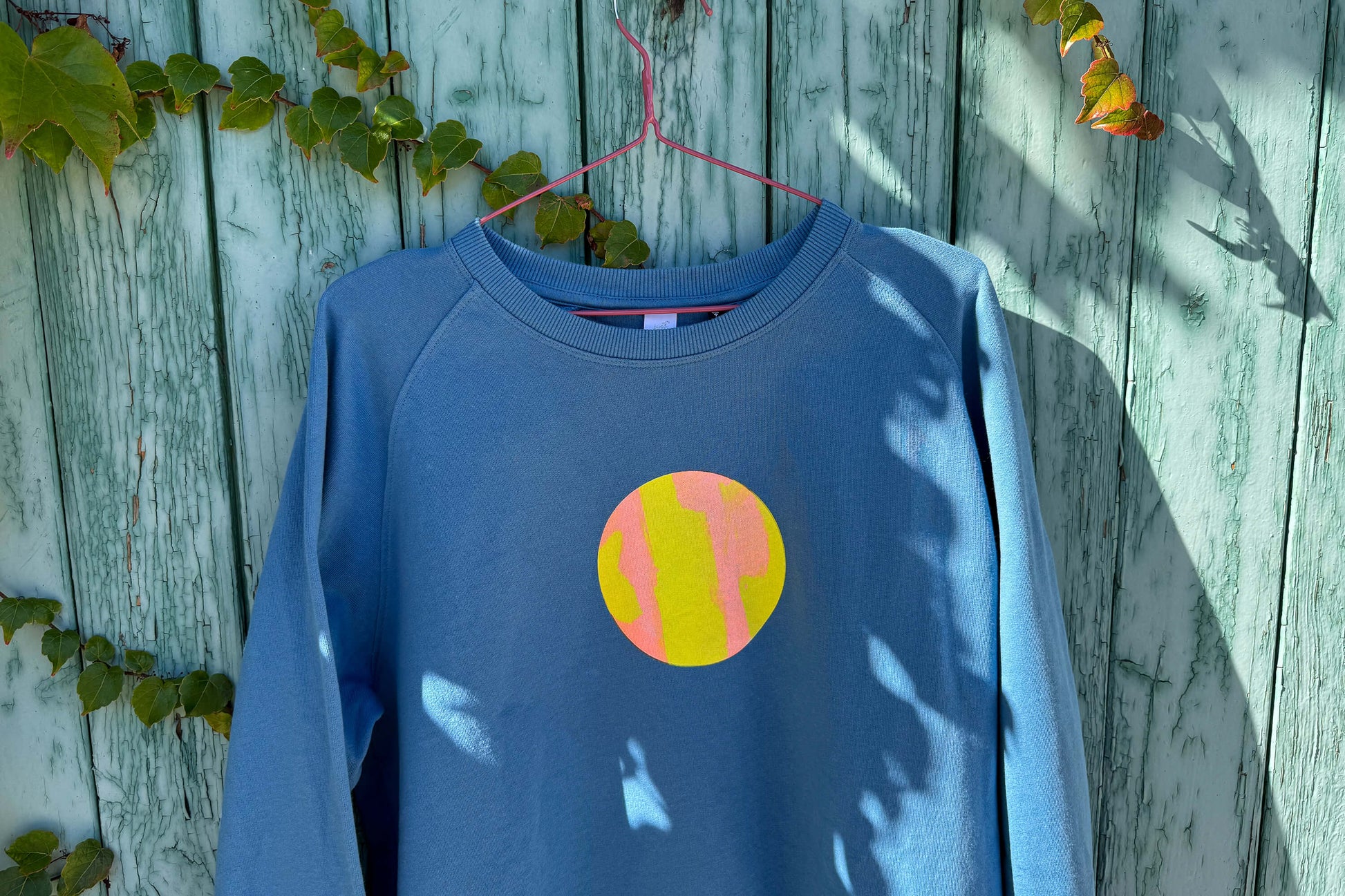 Detailansicht von blauem Sweatshirt vor blauem Hintergrund. Der Pulli hat einen aufgedruckten Kreis in gelb-orange.