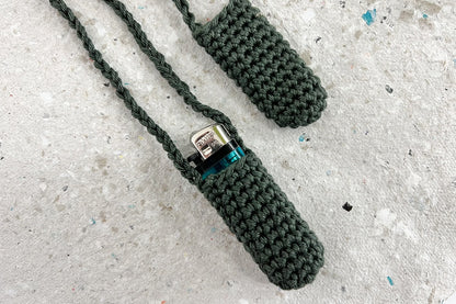 Zwei gehäkelte Taschen fürs Feuerzeug zum Umhängen in olivgrün. Eine Tasche trägt ein blaues Feuerzeug, die andere Tasche ist leer. 