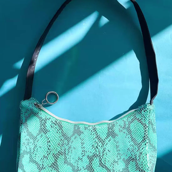 Video: Gesamtansicht blaue Schultertasche in Schlangeleder Optik vor blauem Hintergrund mit schwarzem Gurtband. Hergestellt aus upcycled Materialien von Jeff Every Piece A Friend.
